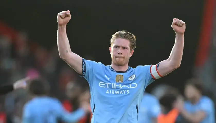 Kevin De Bruyne w jasnoniebieskiej koszulce z podniesionymi rękami, świętujący zwycięstwo.